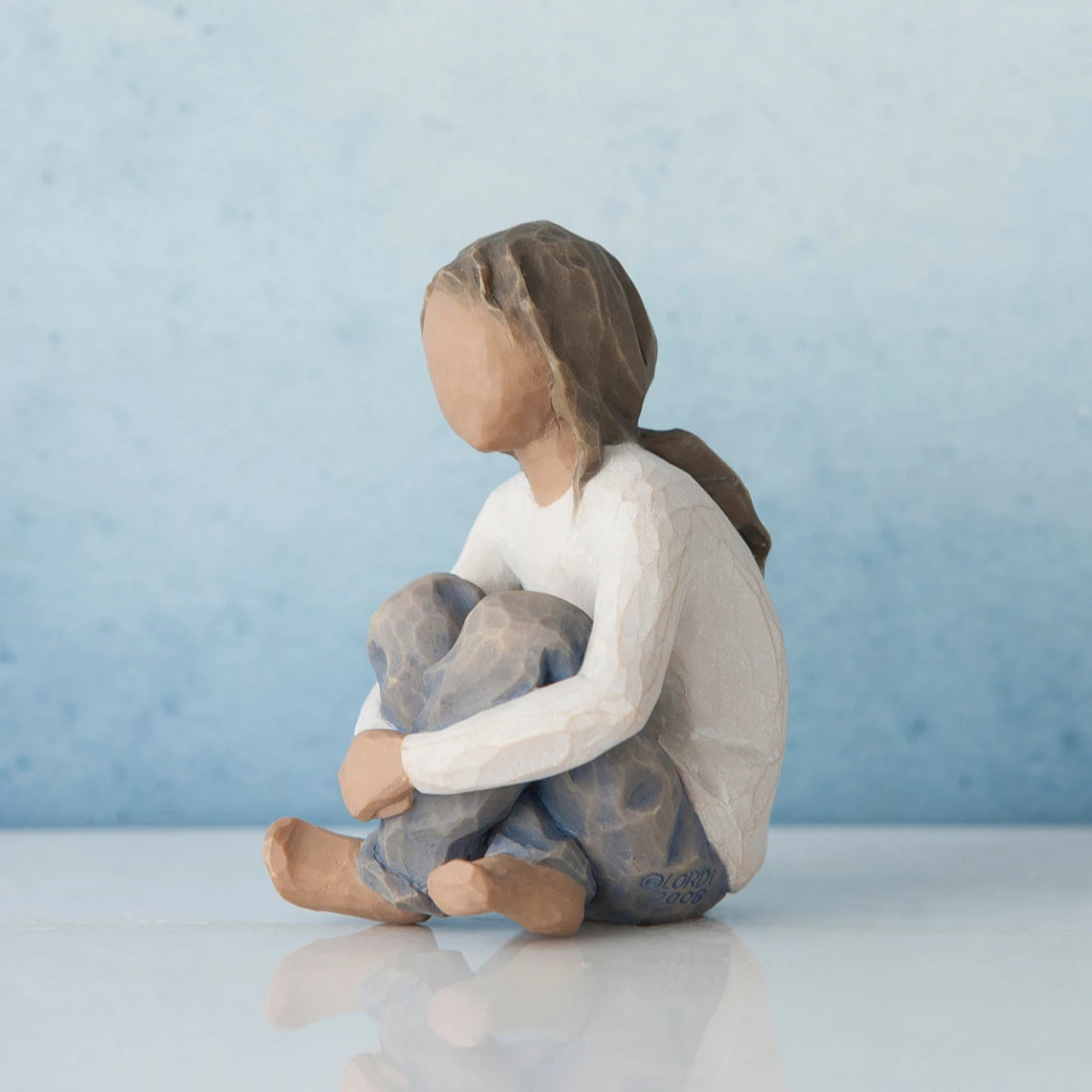 Brunette girl figurine sitting in white shirt and blue jeans with her arms folded on her legs - blue background