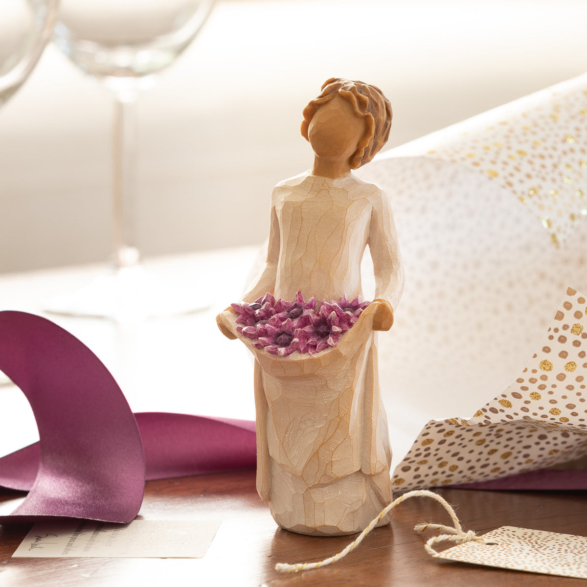 Willow Tree Simple Joys, 5.5”h hand-painted standing figure in cream dress, holding up skirt filled with purple daisies, on table with purple ribbon