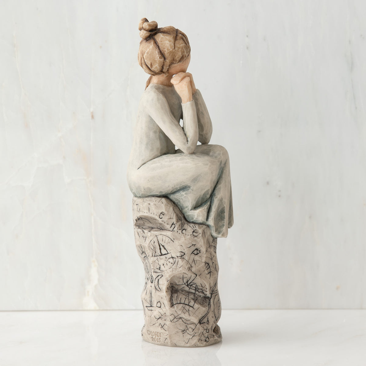 Brunette girl figurine sitting on grey  rock in grey dress with her hands up to her face