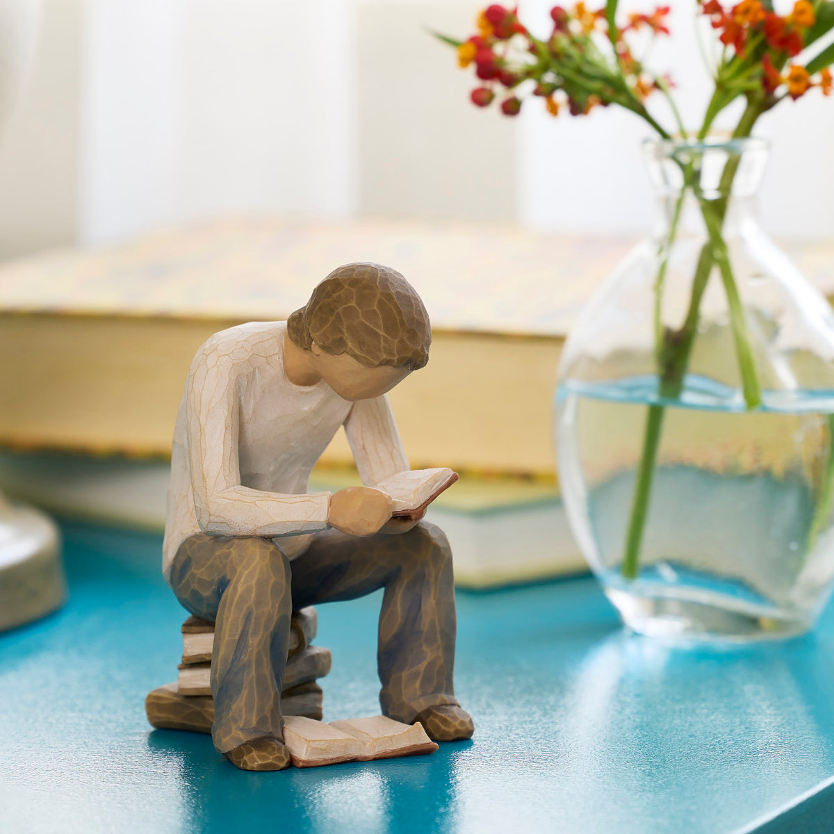 Willow Tree Quest, 4”h hand-painted resin figure of young man in neutral clothes, seated on stack of books and reading, on blue table with books