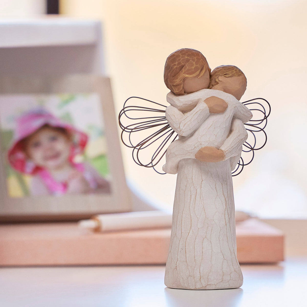 Willow Tree Angel's Embrace, 5"h hand-painted standing resin figure in cream dress with wire wings, holding baby in arms, on shelf with photographs