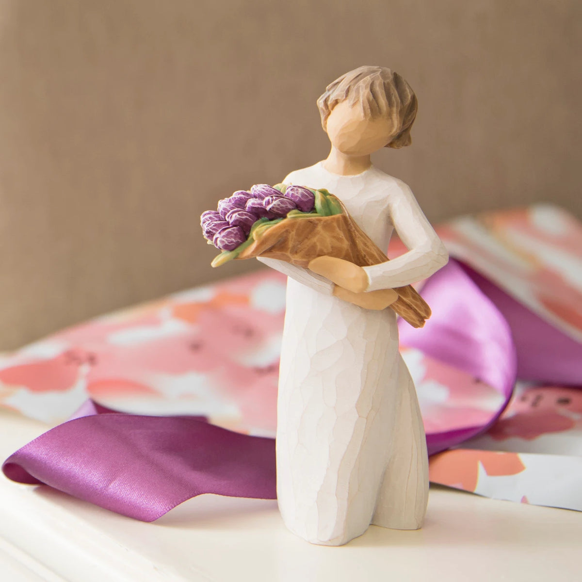 Willow Tree Surprise, 5”h hand-painted standing resin figure in cream dress, holding bouquet of purple tulips, on desk with gift wrap