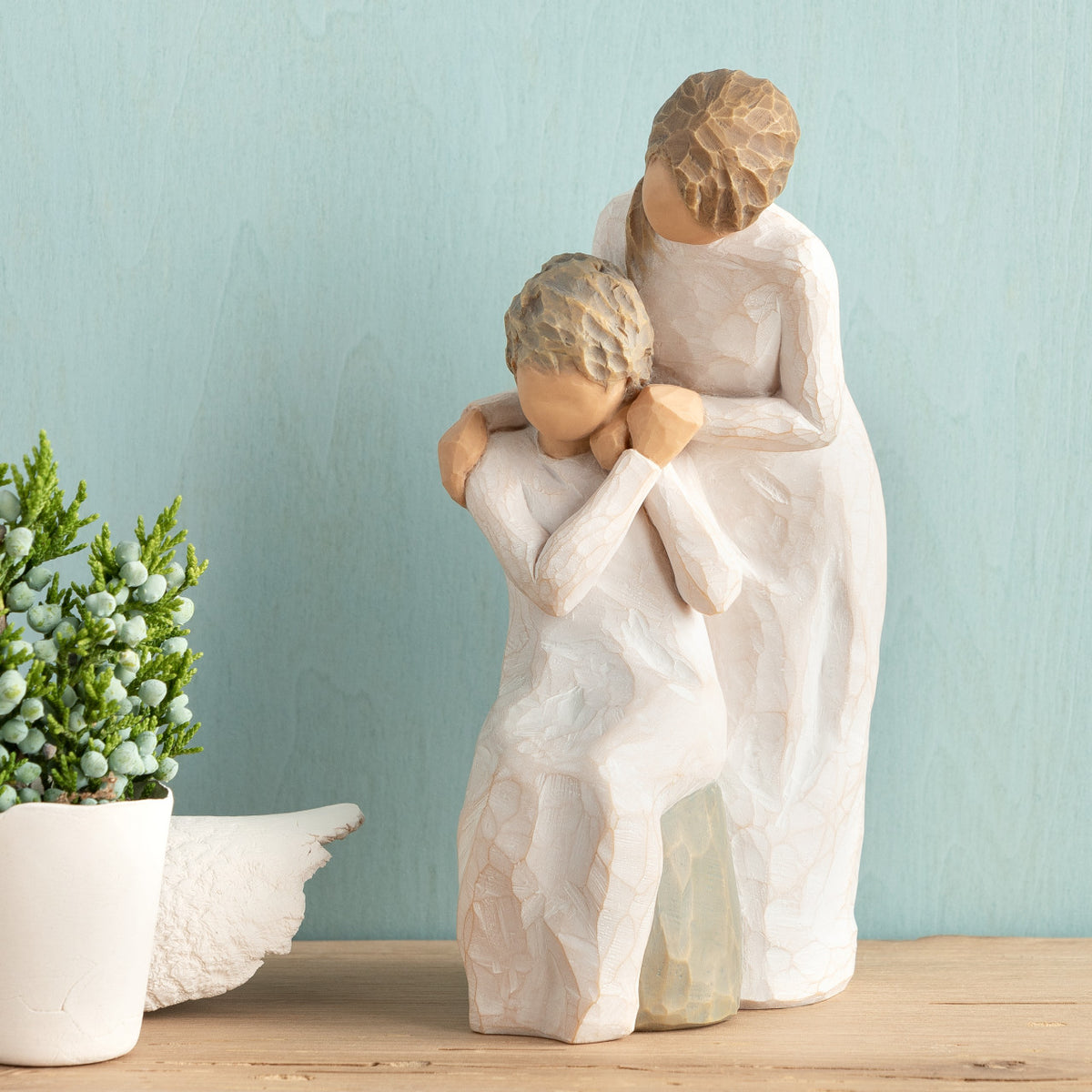 Willow Tree Loving My Mother, 6.5”h hand-painted resin figure of seated older woman with younger woman standing at her shoulder, both in cream dresses, hands clasped, in front of muted teal painting