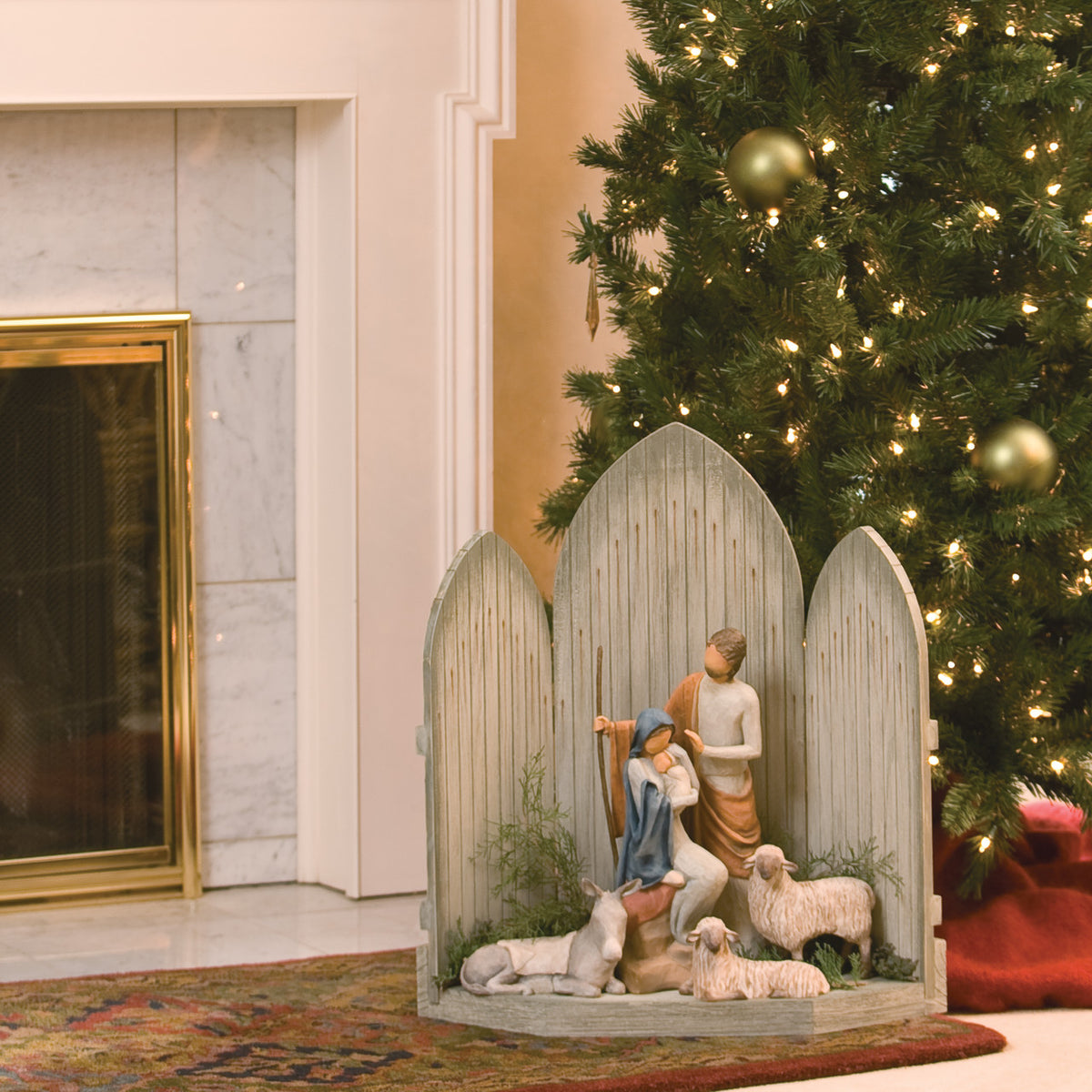 Far view of light grey barn figurine tri fold with mary and joesph figurines around two white sheep and one donkey - christmas tree in the background
