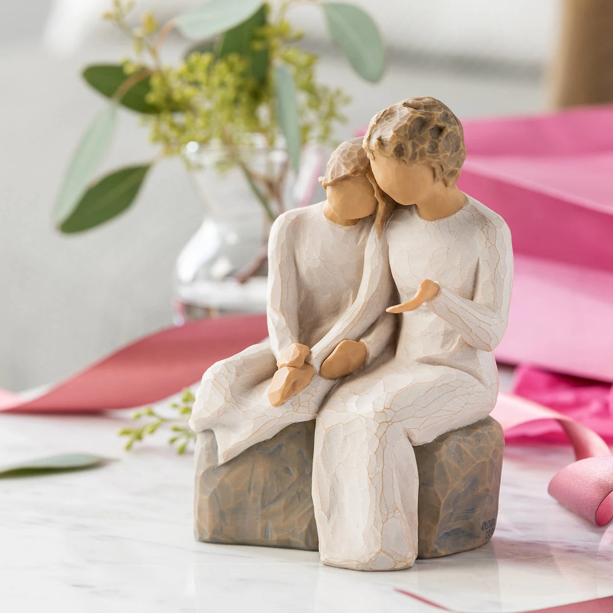 Willow Tree With my Grandmother, 5.5" hand-painted resin figure of older woman seated on gray bench, holding hands with young girl, both in cream dresses, on table with pink wrapping