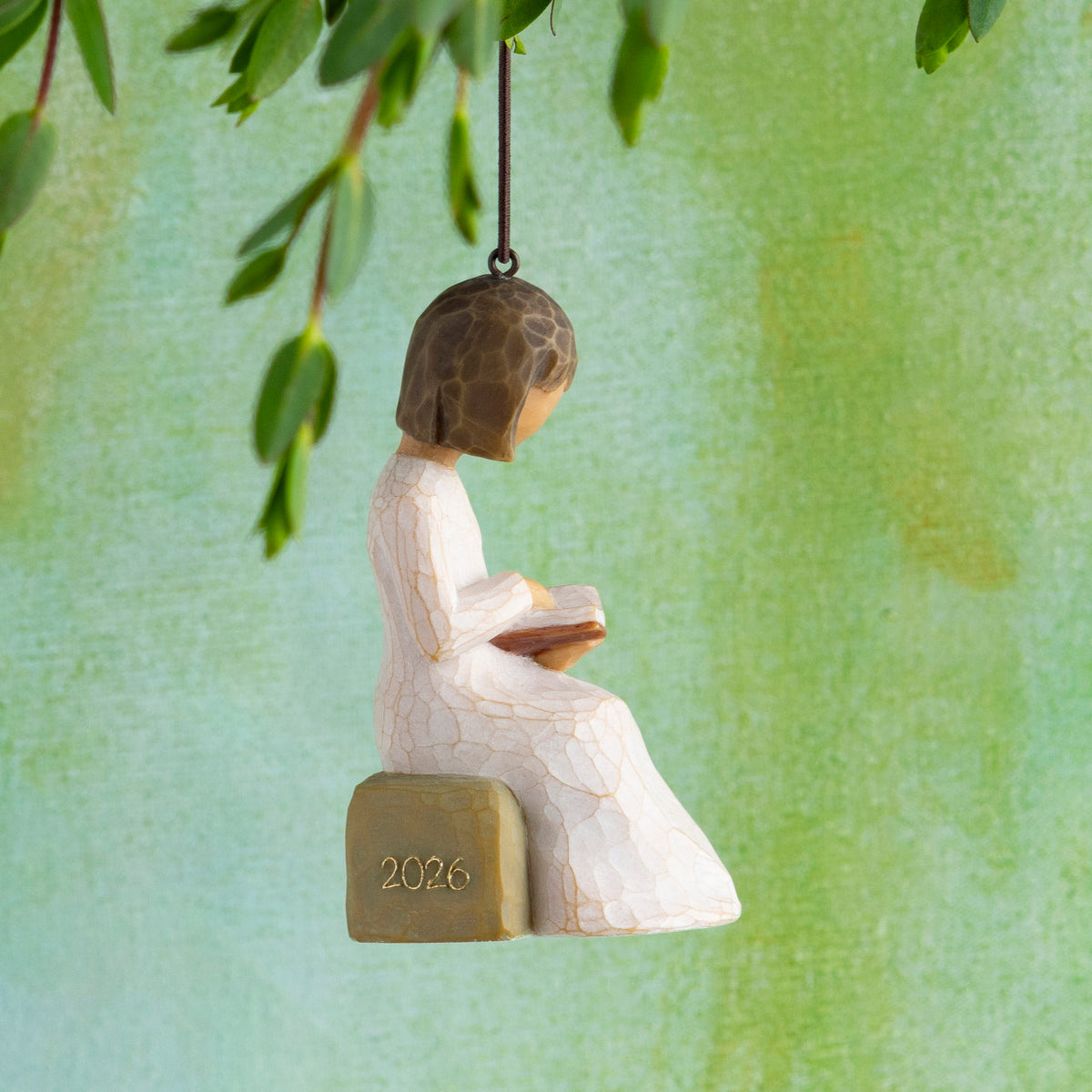 Willow Tree 2026 Ornament, 3”h hand-painted resin figure in cream dress, reading book, seated on rock with 2026 written in gold, brown elastic cord, hanging in front of green painting
