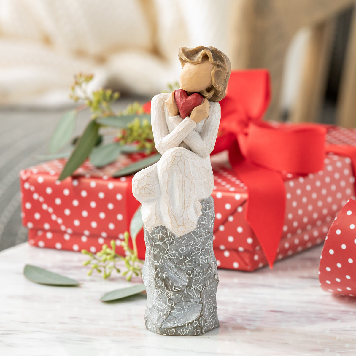 Willow Tree Always, 6”h hand-painted figure in cream dress, knees drawn up to chest, holding red heart to face, seated on gray rock etched with symbols of love, on table with gifts