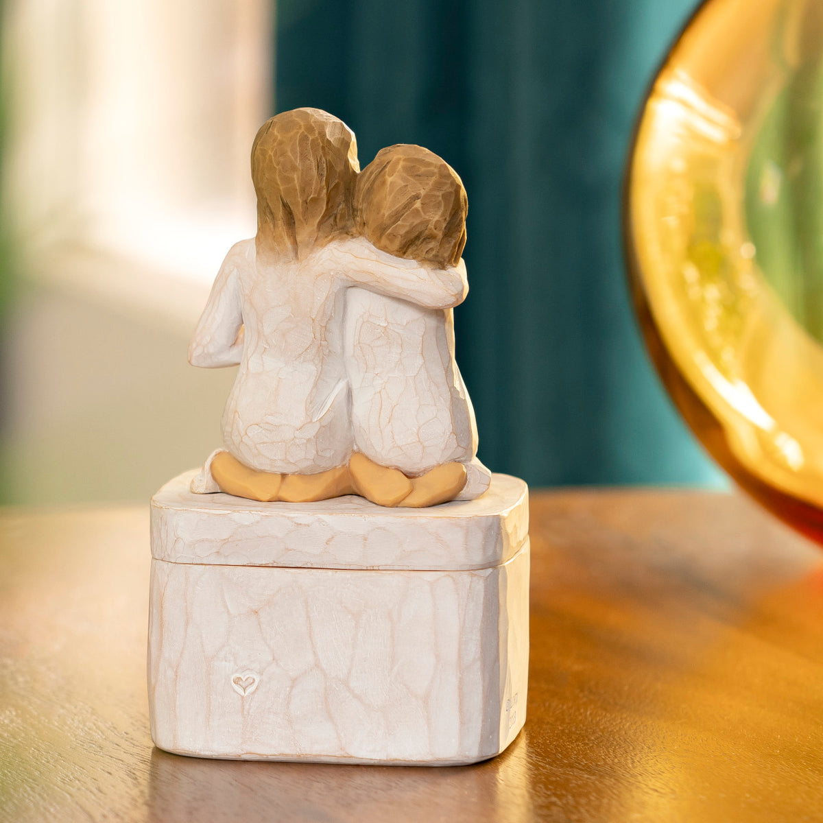 Willow Tree Sister mine Keepsake Box, back view of 3"sq hand-painted resin box with 3.5”H figure of two kneeling girls in cream dresses, arms around each other, affixed to lid, on wooden table
