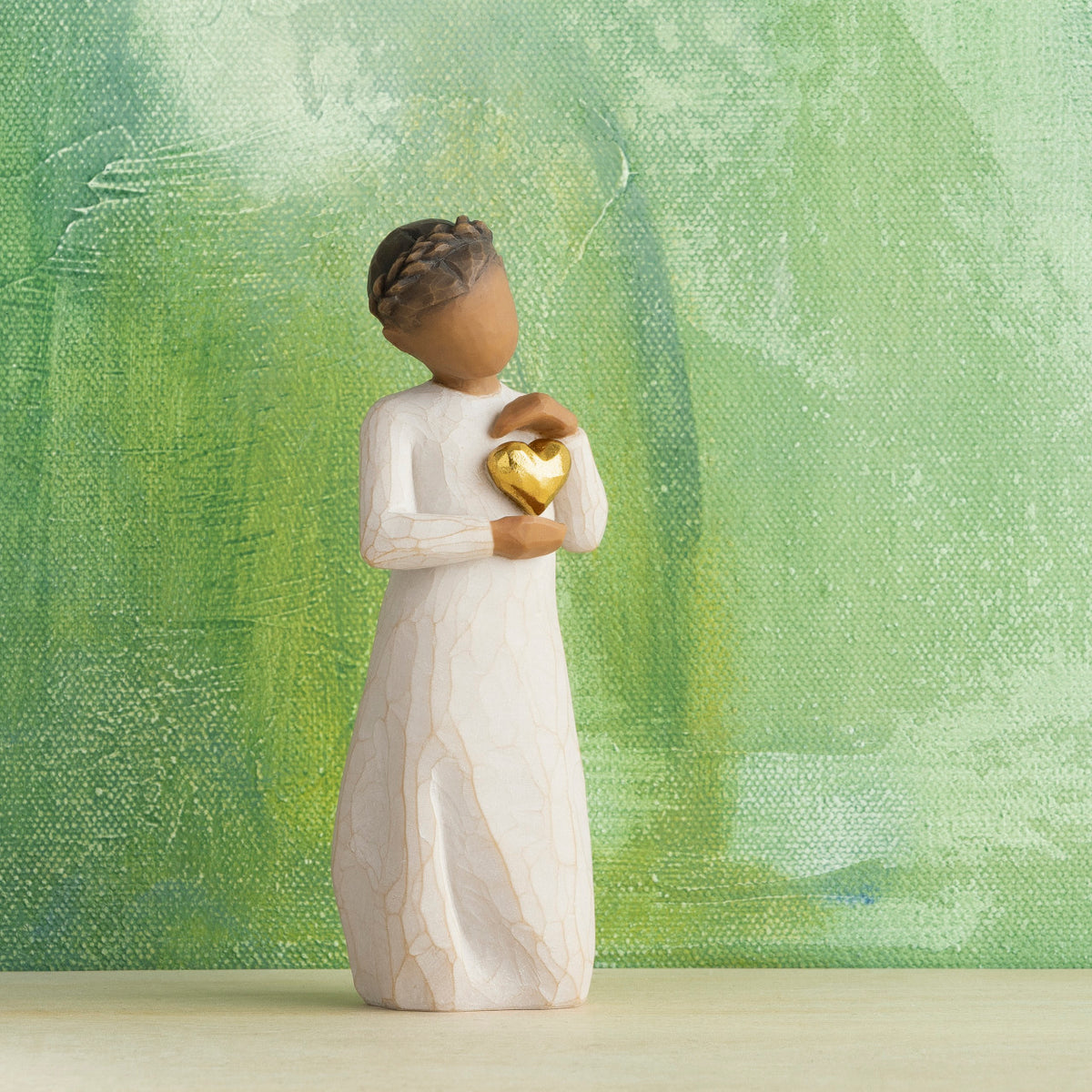 Willow Tree Keepsake, 5.5”h hand-painted standing resin figure with darker skin and hair, in cream dress, framing gold heart with both hands on her chest, in front of green painting