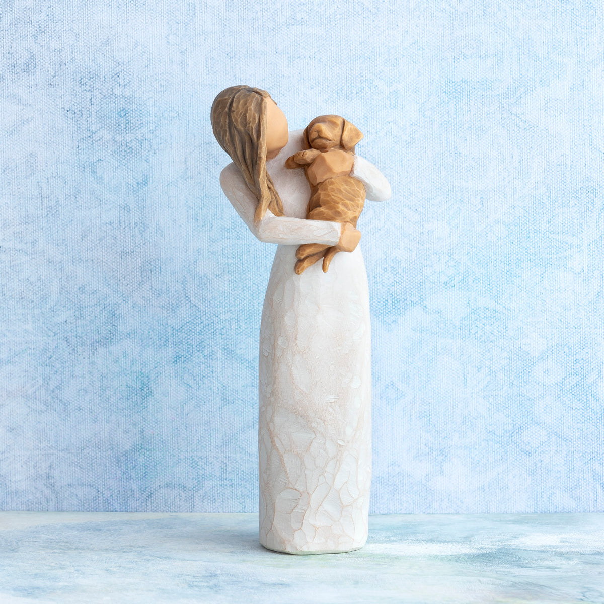Standing female figure in cream dress, holding golden color puppy in her arms, on blue painted background