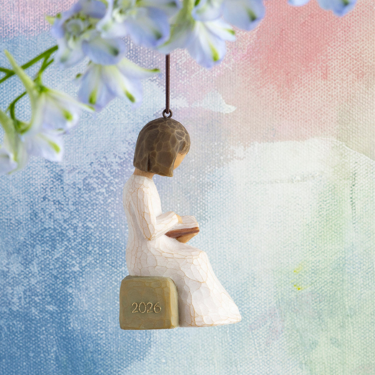Willow Tree 2026 Ornament, 3”h hand-painted resin figure in cream dress, reading book, seated on rock with 2026 written in gold, brown elastic cord, hanging in front of multi-pastel painting