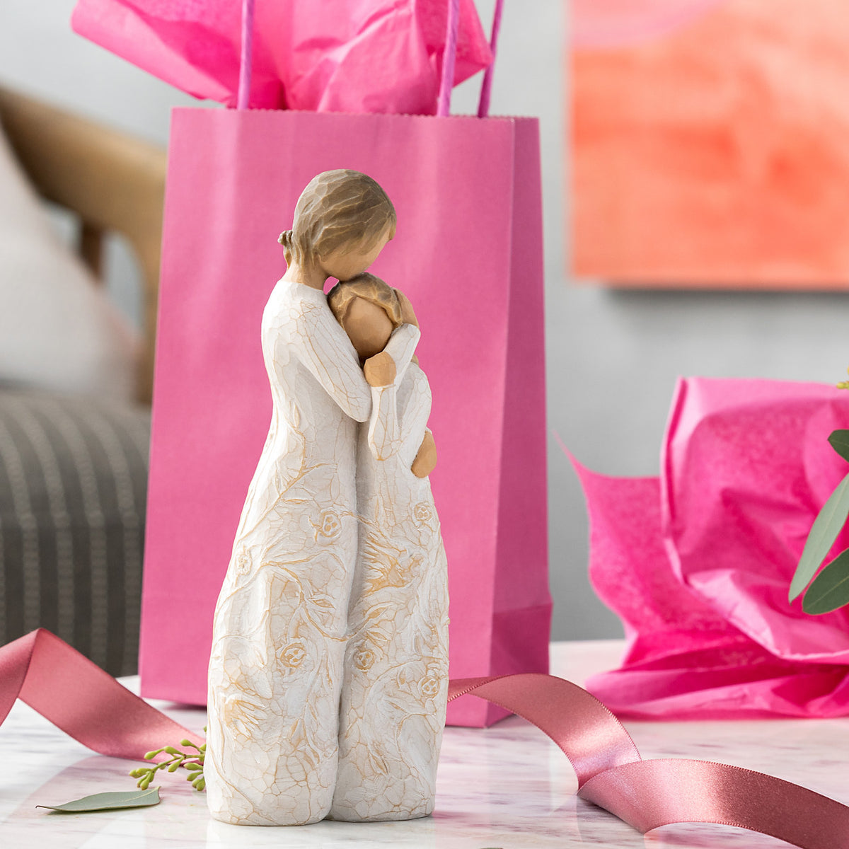 Willow Tree Close to me, 8"h hand-painted standing resin figure of woman embracing tween girl, both in cream dresses carved with intertwined vines and leaves, on table with pink gift bag