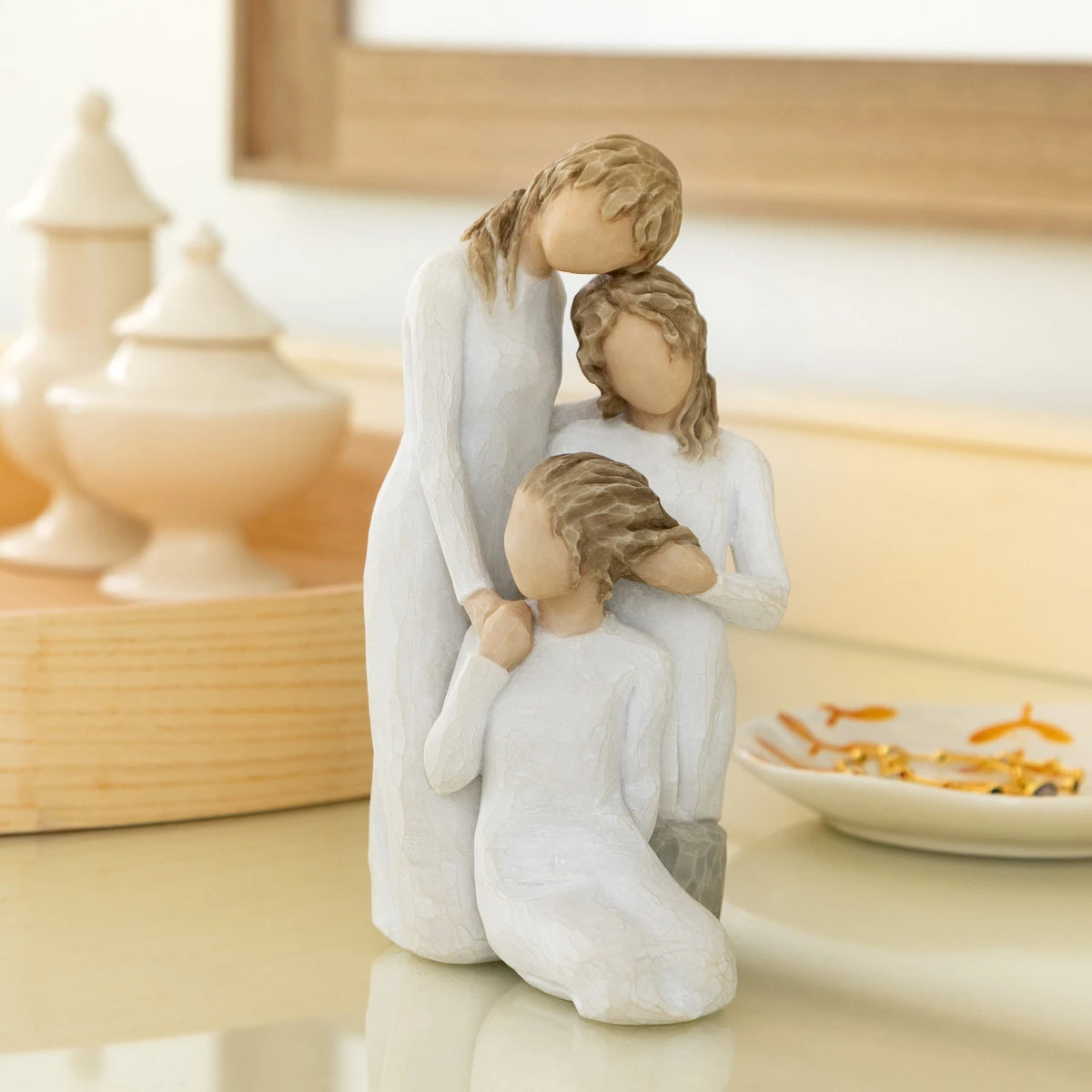 Willow Tree Our healing touch, 6.5”h hand-painted resin figure of three women; one standing with arm around kneeling girl, holding hand of seated girl, kneeling girl arranging hair of seated girl, all in cream dresses, on bedside table