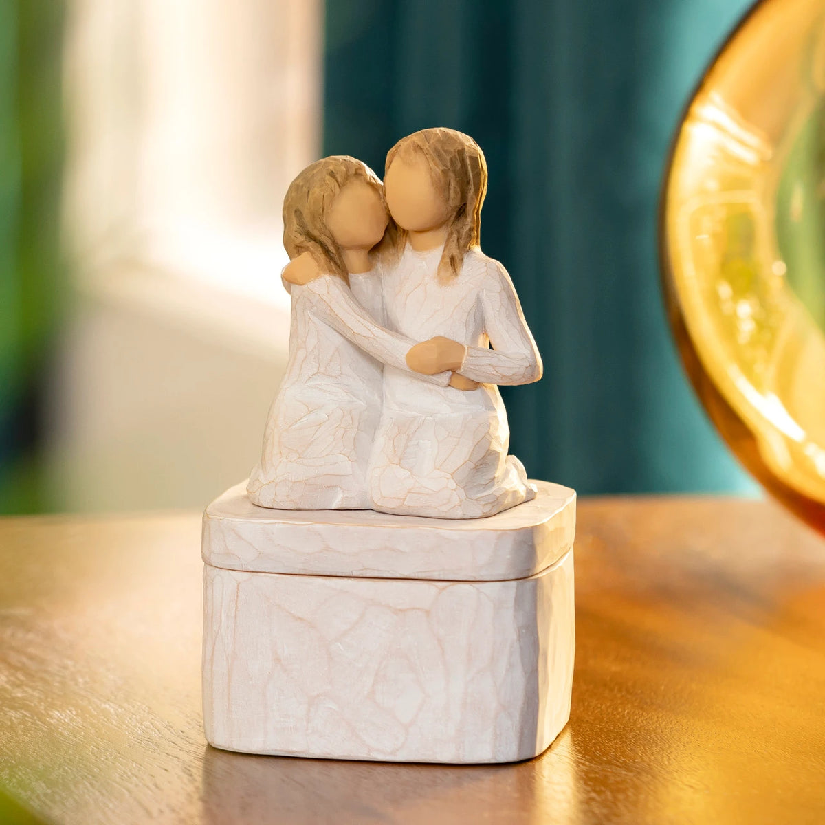 Willow Tree Sister mine Keepsake Box, front view of 3"sq hand-painted resin box with 3.5”H figure of two kneeling girls in cream dresses, arms around each other, affixed to lid, on wooden table