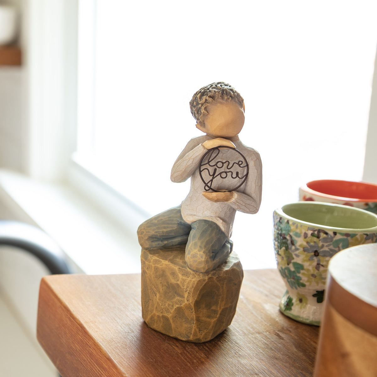 Willow Tree Love you too (lighter skin), 4.5”h hand-painted resin figure of young child in cream shirt and jeans, kneeling on rock, holding wire circle with ‘love you’ inside, on shelf by window