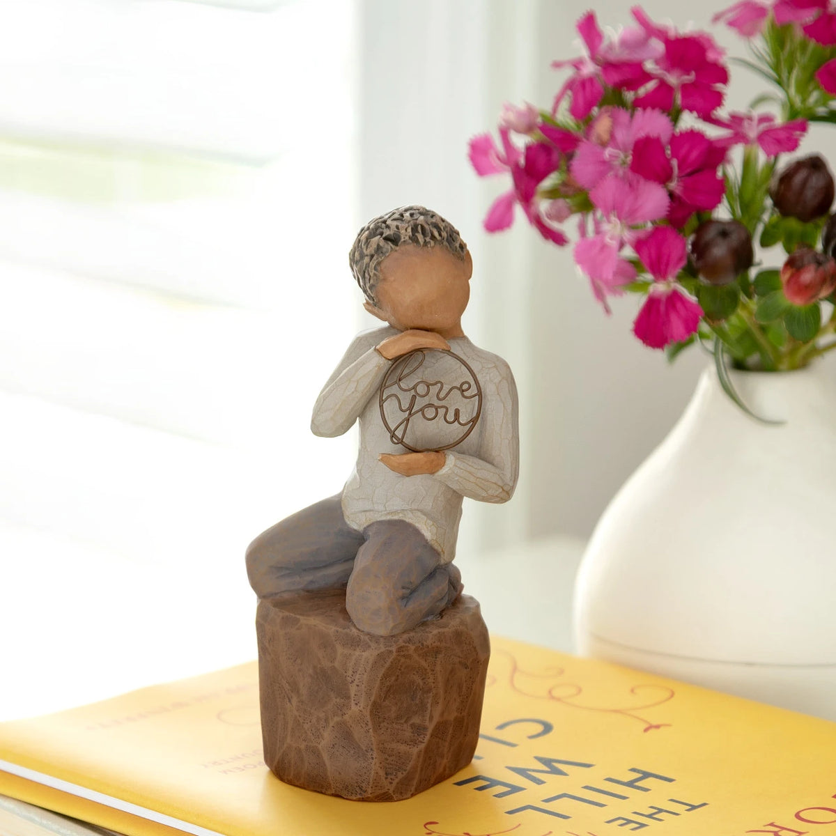 Willow Tree Love you too (darker skin), 4.5”h hand-painted resin figure of young child in cream shirt and jeans, kneeling on rock, holding wire circle with ‘love you’ inside, on shelf by window