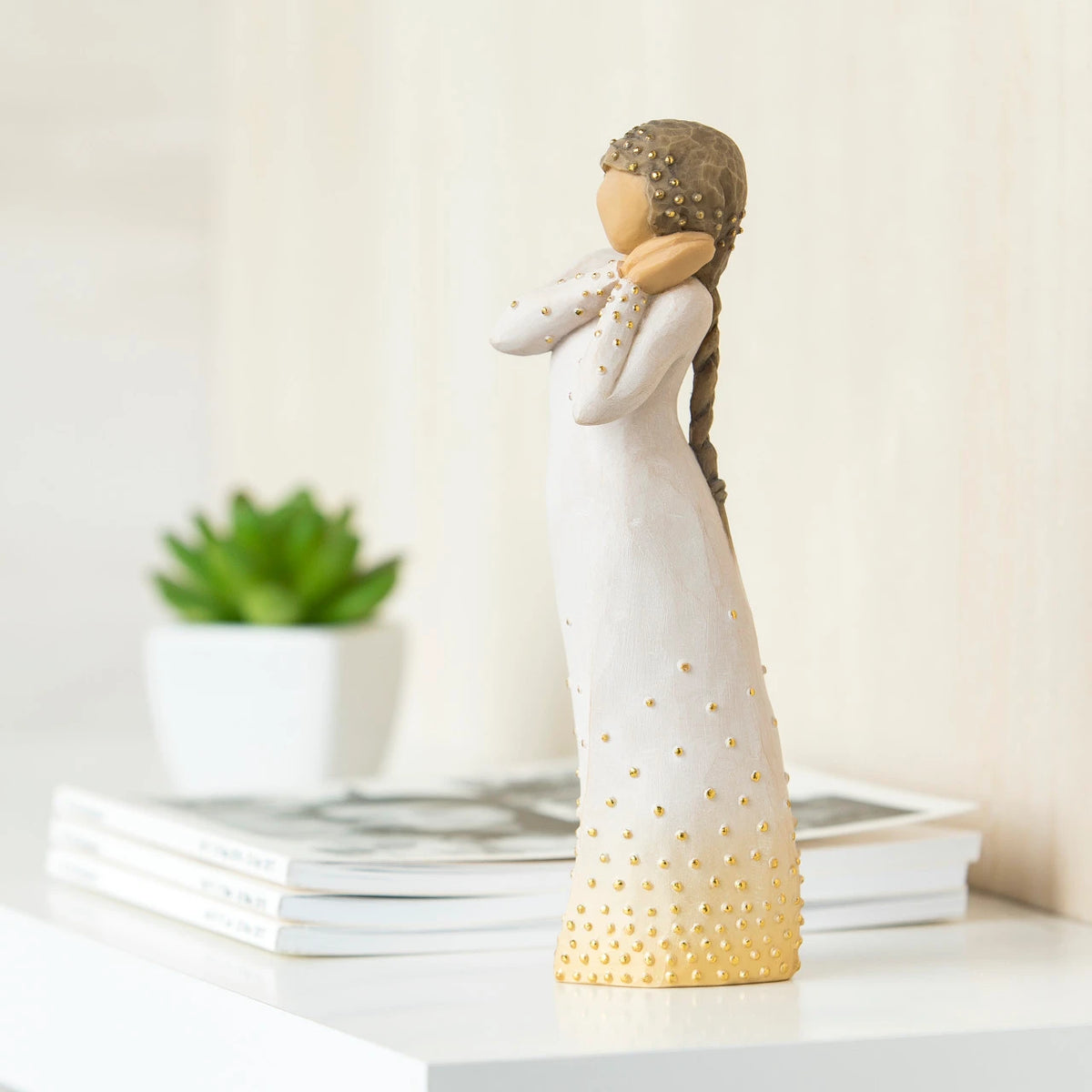 Willow Tree Wishing, 6.5”h hand-painted standing resin figure holding hands to face in wistful gesture, bottom of cream dress has gold-leaf dots, on living room table